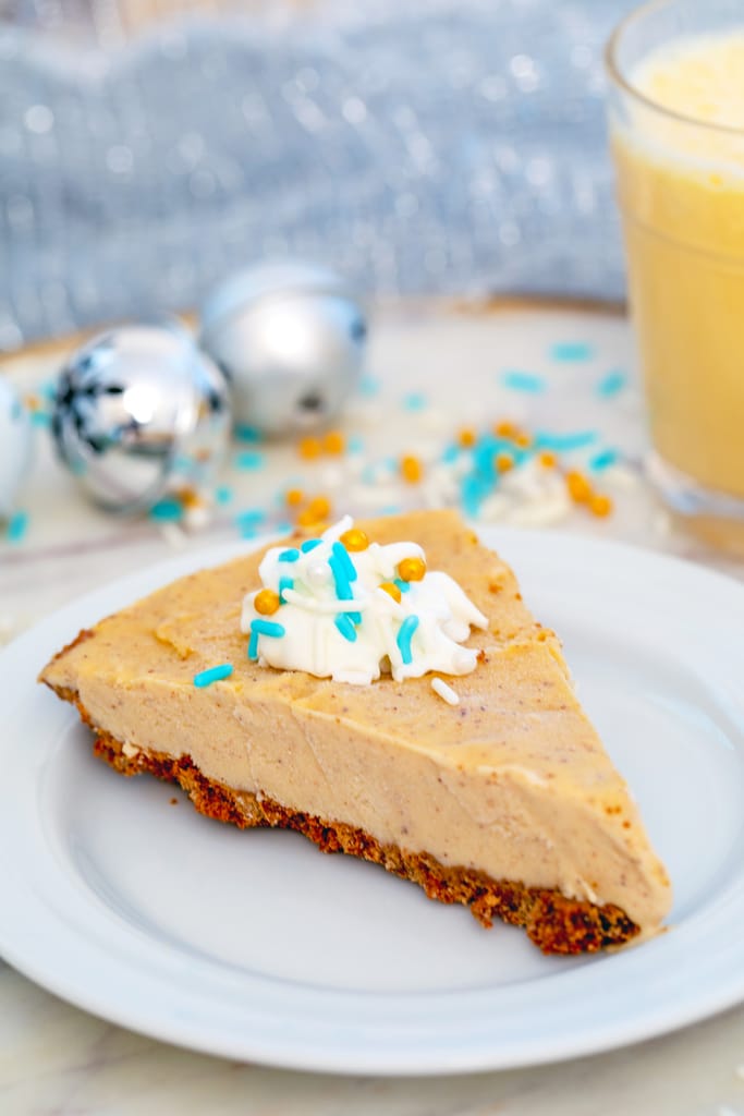 View of a slice of eggnog ice cream pie on a white plate with a dollop of rum whipped cream and blue and gold sprinkles with Christmas bells, sprinkles, and a glass of eggnog in the background.
