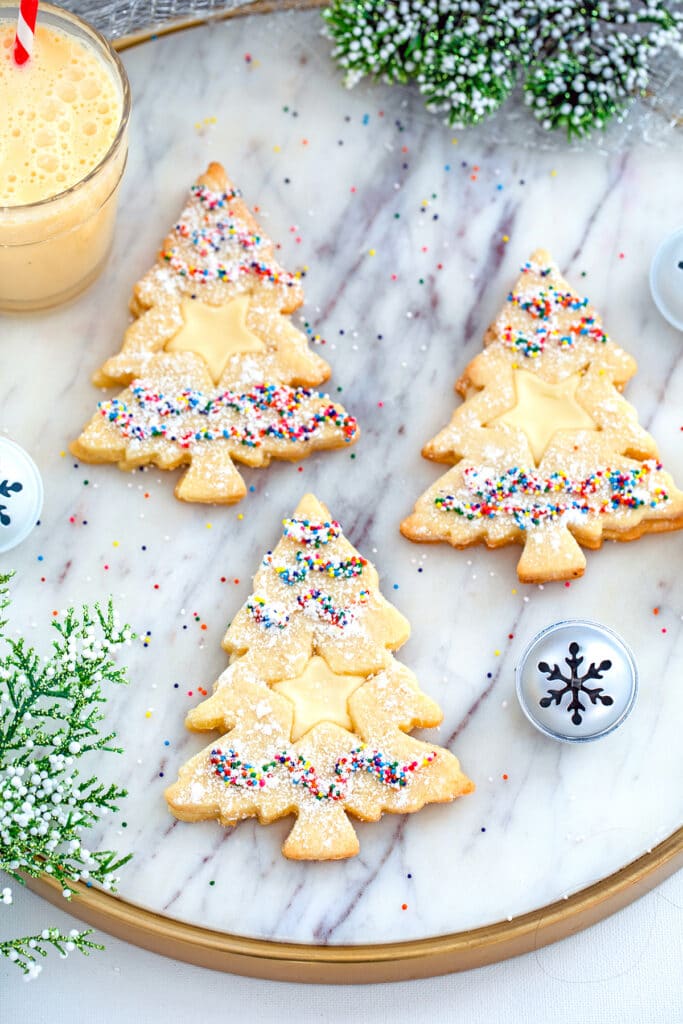 Overhead view of three Christmas tree-shaped eggnog linzer cookies with star cutouts and sprinkles on a marbled surface with glass of eggnog, holly, and Christmas bells in the background