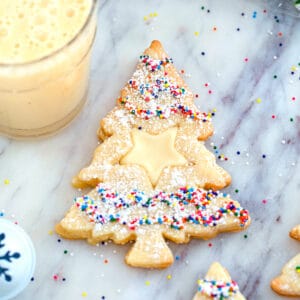 Overhead close-up view of Christmas tree-shaped eggnog linzer cookies with a star cutout and sprinkles on a marble surface with glass of eggnog in the background