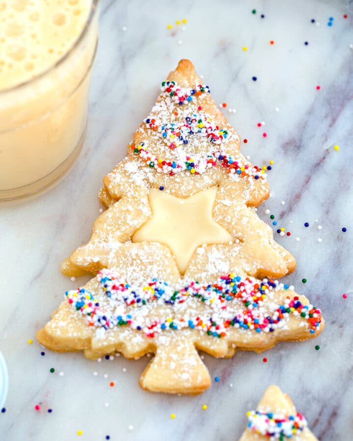 Overhead close-up view of Christmas tree-shaped eggnog linzer cookies with a star cutout and sprinkles on a marble surface with glass of eggnog in the background