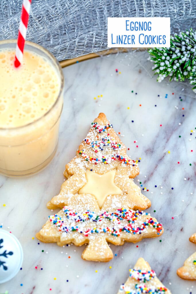 Overhead close-up view of Christmas tree-shaped eggnog linzer cookies with a star cutout and sprinkles on a marble surface with glass of eggnog in the background and recipe title at top