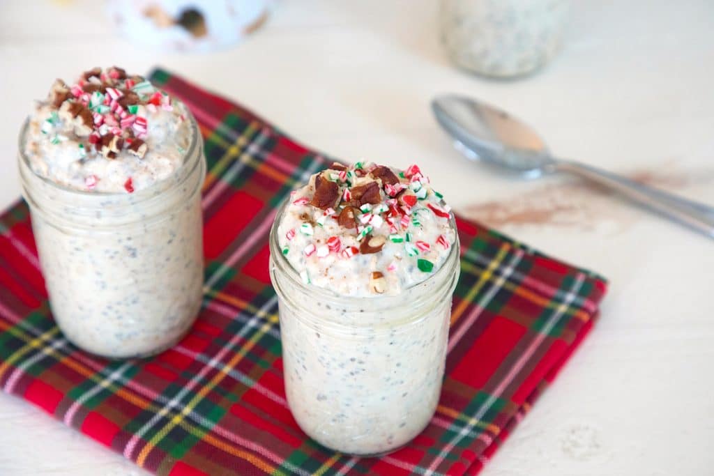 Overhead landscape view of two mason jars of eggnog overnight oats topped with crushed candy bars and pecans with spoon in the background