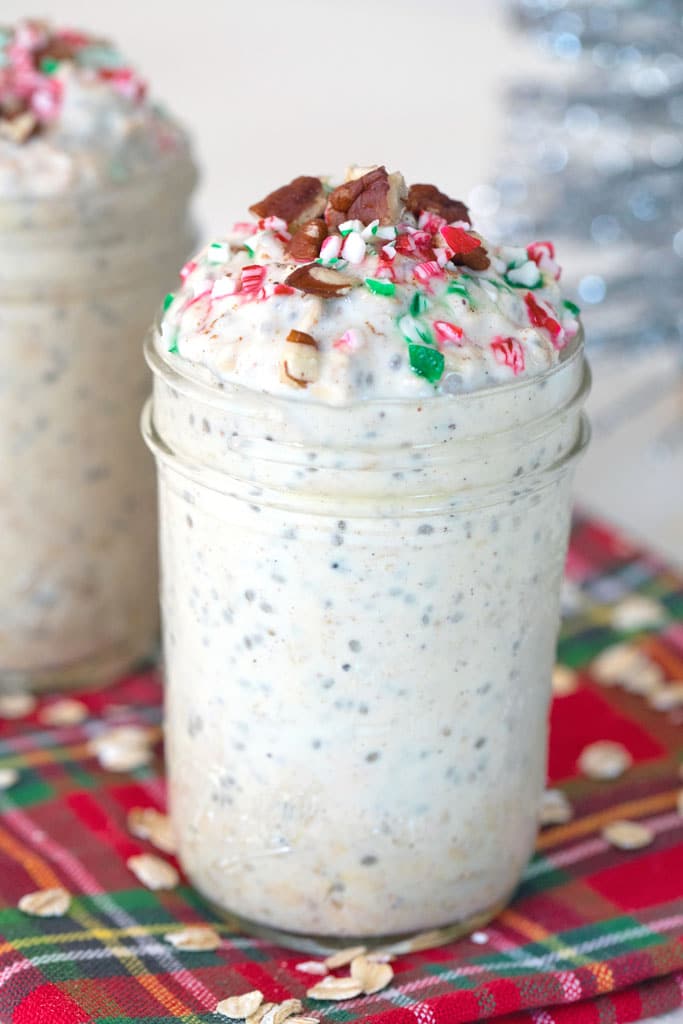 Head-on view of a mason jar filled with eggnog overnight oats topped with crushed candy canes and pecans with second jar and silver tinsel in the background