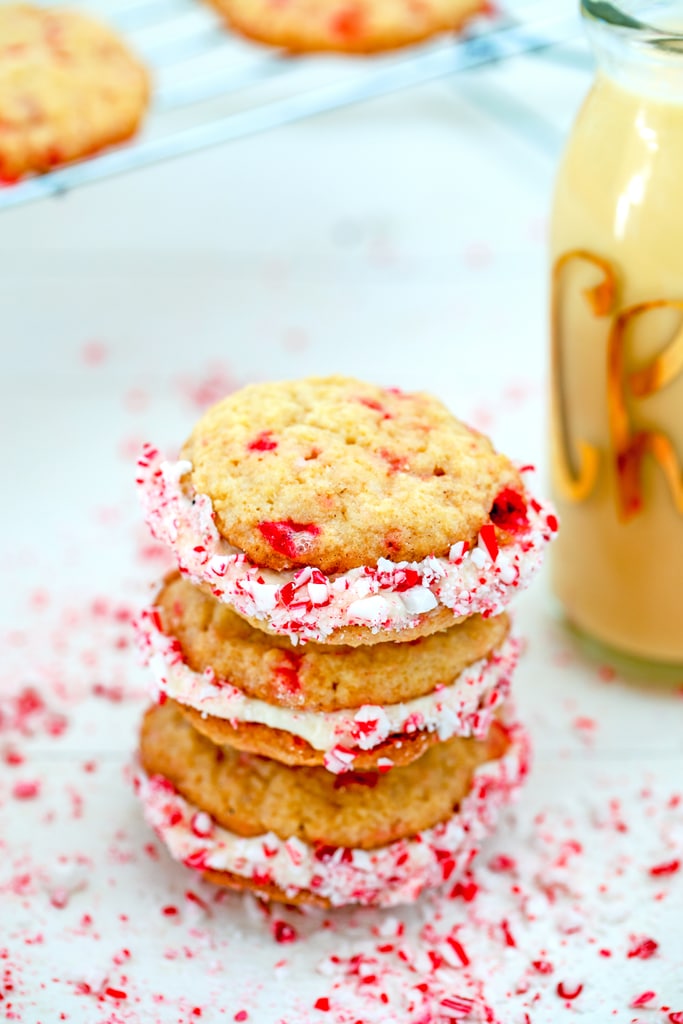 Overhead view of three eggnog peppermint sandwich cookies stacked on each other with crushed candy canes, glass of eggnog, and baking rack with more cookies in the background