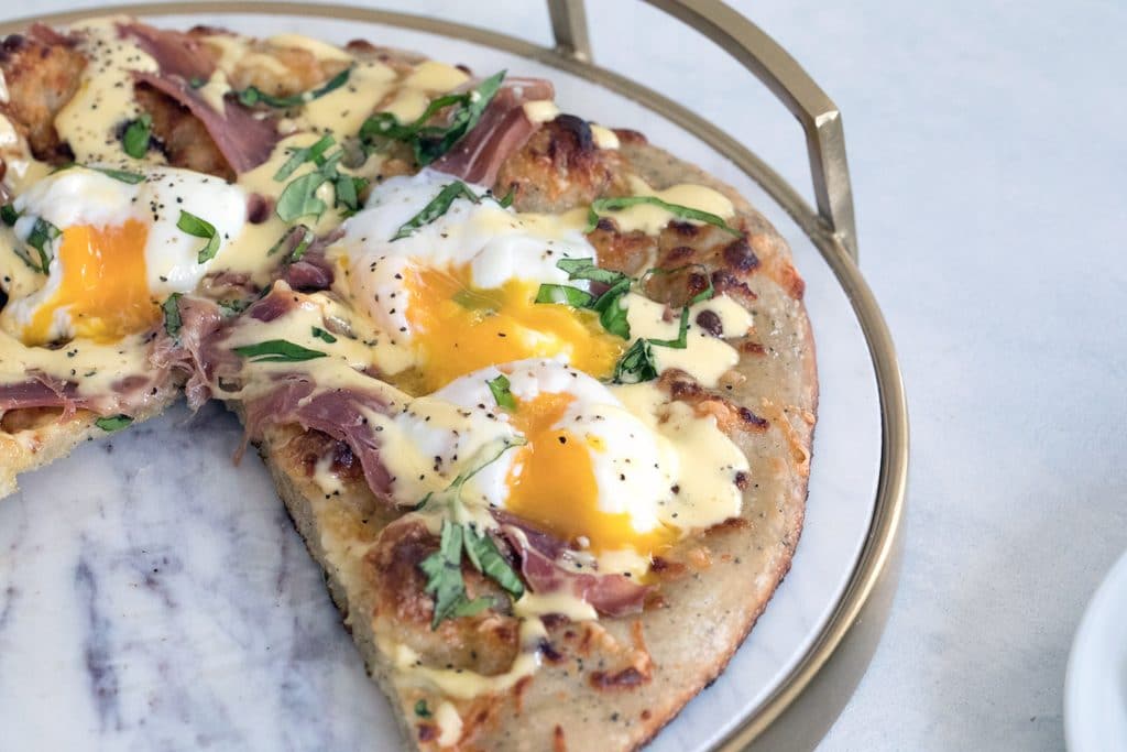 Landscape view of eggs benedict pizza on a marble tray with runny eggs, prosciutto, and hollandaise sauce over the top