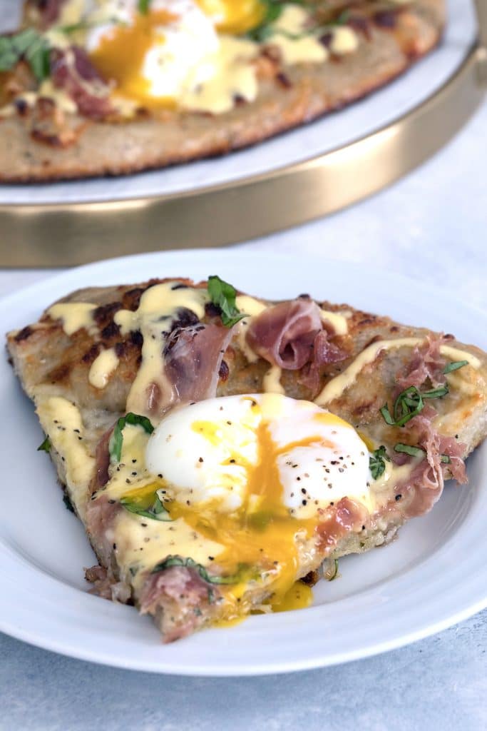 Head-on view of a slice of eggs benedict pizza on a white plate with runny egg, prosciutto, and hollandaise sauce