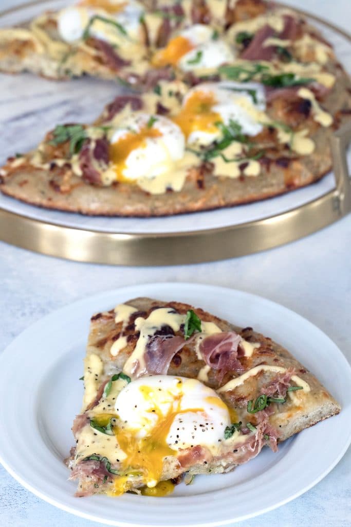 Overhead view of a slice of eggs benedict pizza with runny egg on a white plate with rest of brunch pizza in background