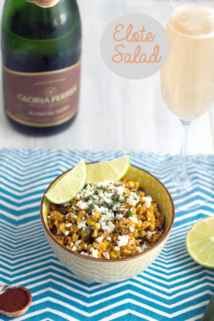 Head-on view of elote salad on a blue striped towel with lim garnish and bottle and glass of champagne in the background with "Elote Salad" text at top
