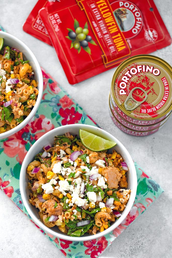 Bird's eye view of two bowls of elote tuna salad on a flowered towel with tuna cans and pouches in the background