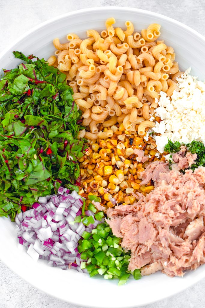Overhead view of elote tuna salad ingredients in a serving bowl, including tuna, jalapeño, red onion, chard, macaroni, cotija cheese, cilantro, and grilled corn