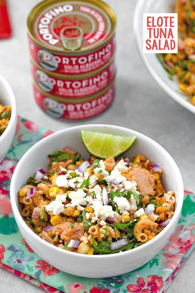 Head-on view of a white bowl of elote tuna salad with lime wedge garnish and Portofino Tuna cans and a serving bowl of salad in the background with recipe title at top