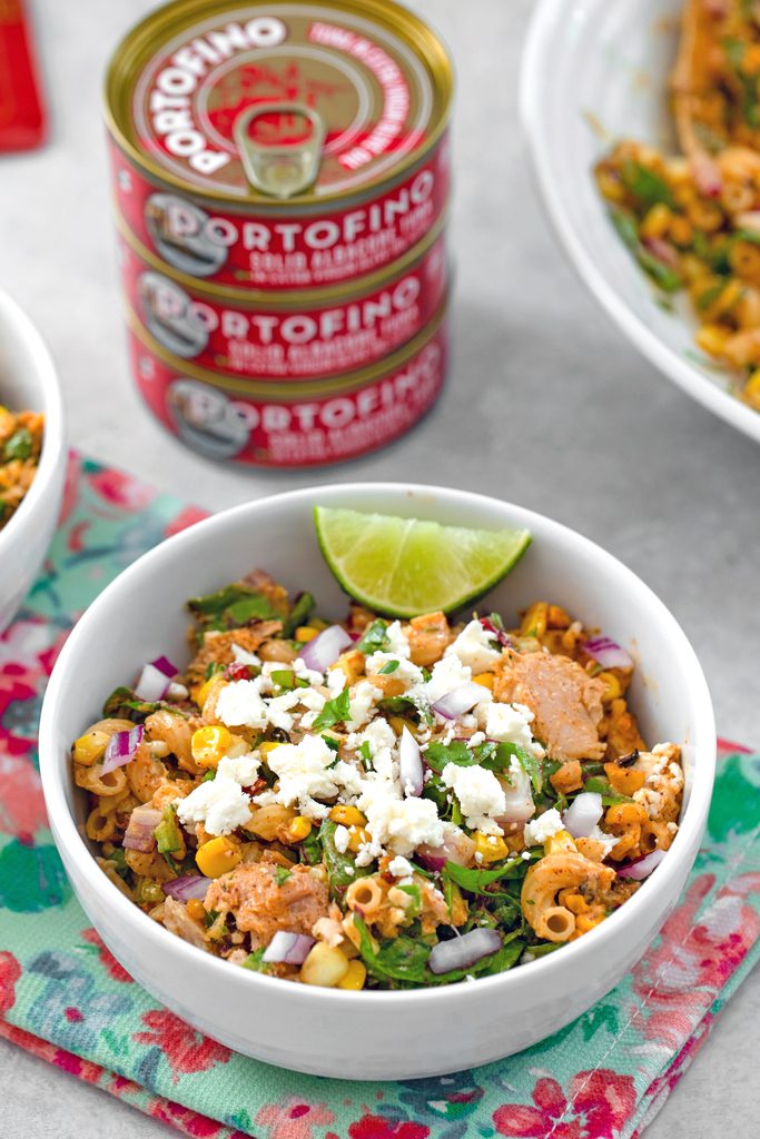 Head-on view of a white bowl of elote tuna salad with lime wedge garnish and Portofino Tuna cans and a serving bowl of salad in the background