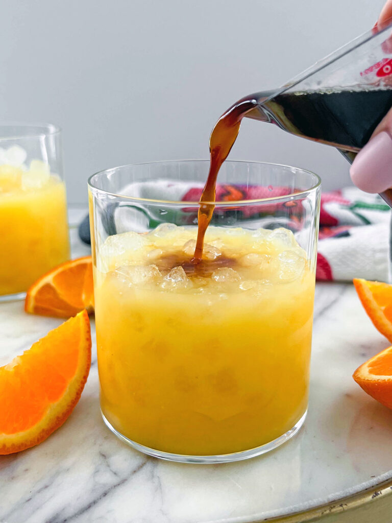 Espresso being poured into a glass of orange juice with orange slices all around.