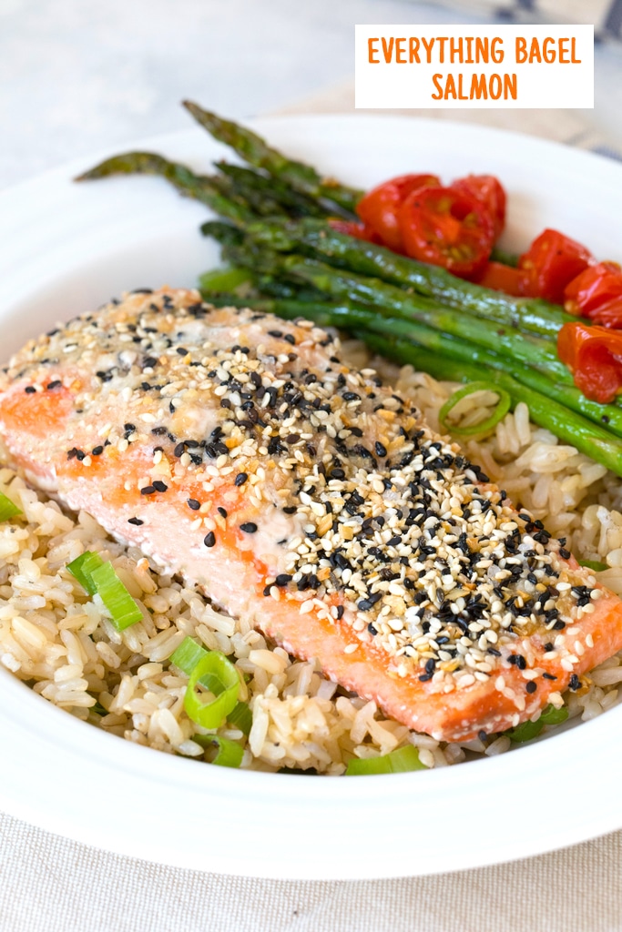 Head-on view of a piece of salmon coated with everything bagel spice over a bed of brown rice with asparagus  and grape tomatoes in the background and recipe title at top