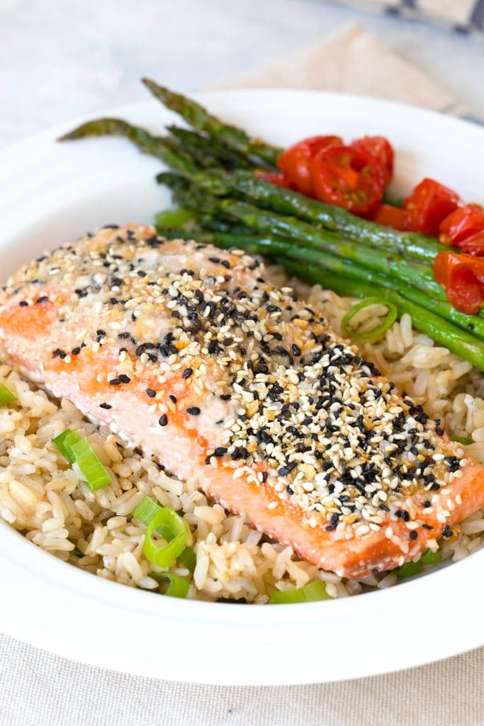 Head-on view of a piece of salmon coated with everything bagel spice over a bed of brown rice with asparagus  and grape tomatoes in the background.