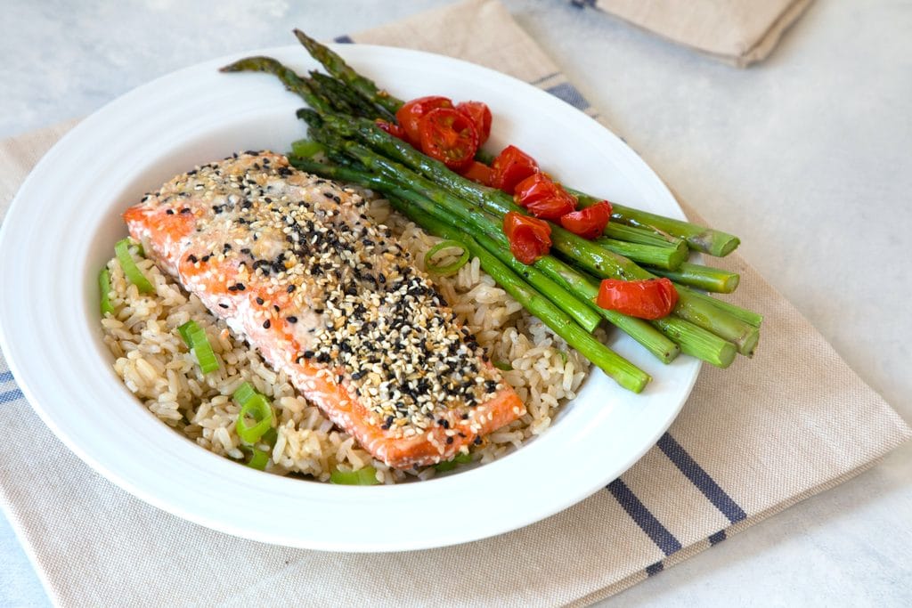 Landscape overhead view of a piece of salmon coated in everything bagel spice over a bed of brown rice with asparagus and grape tomatoes.