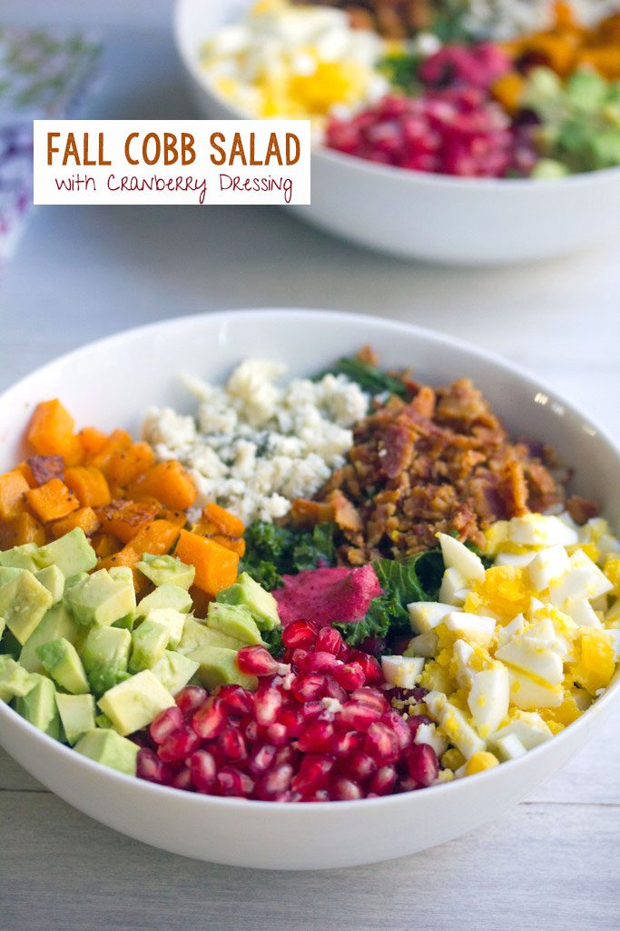 Head-on view of a fall cobb salad with kale, pomegranate, avocado, butternut squash, gorgonzola, bacon, and hardboiled egg with second bowl of salad in the background and recipe title at top