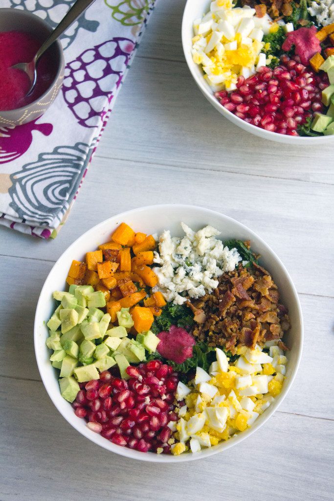 Bird's eye view of two bowls of fall cobb salad with kale, pomegranate, avocado, butternut squash, gorgonzola, bacon, and hardboiled eggs with bowl of cranberry dressing in the background