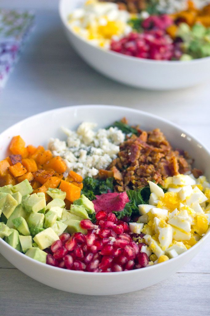 Head-on view of a fall cobb salad with kale, pomegranate, avocado, butternut squash, gorgonzola, bacon, and hardboiled egg with second bowl of salad in the background