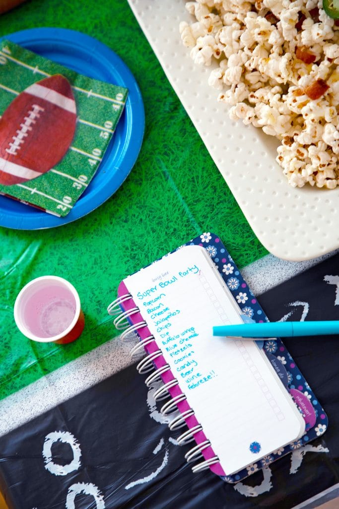 Bird's eye view of Super Bowl to-do list with cups, plates, football napkins, and platter of bacon jalapeño cheddar popcorn on a football field tablecloth