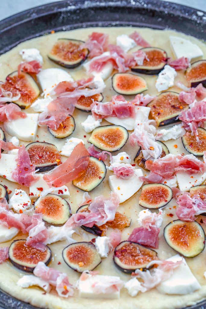 Overhead view of flatbread dough rolled out on pan and topped with sliced figs, prosciutto, goat cheese, and mozzarella