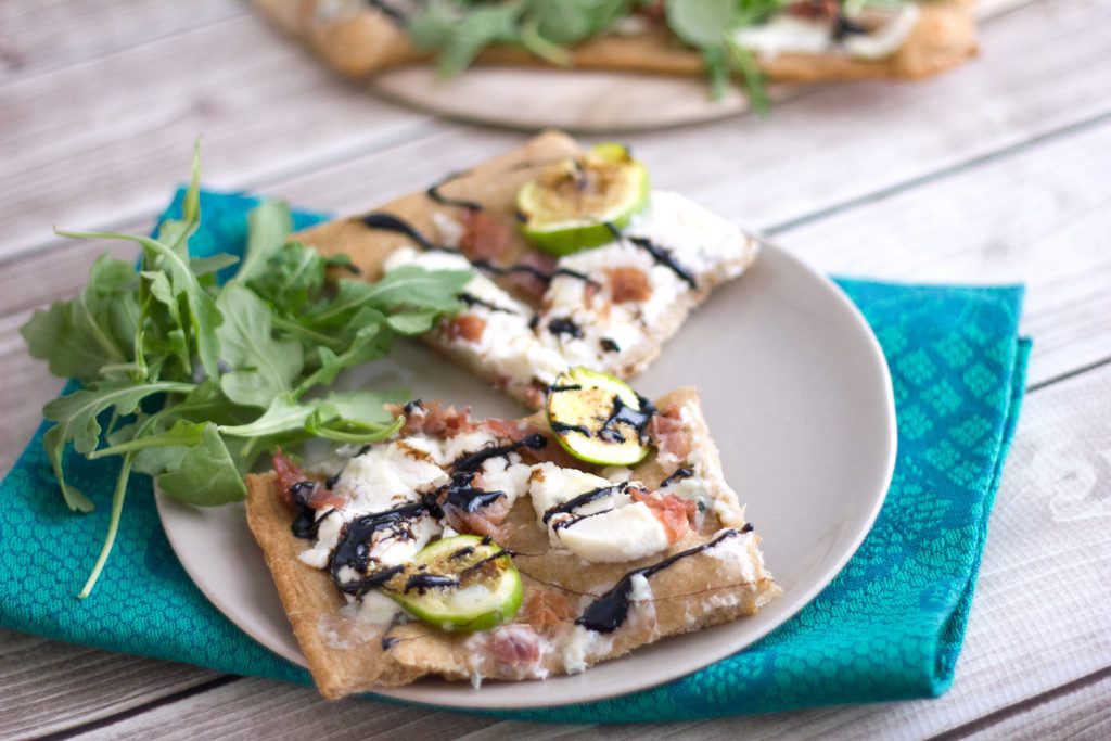 Landscape view of two slices of fig, prosciutto, and ricotta flatbread on a plate with arugula on the side and remaining pizza in the background