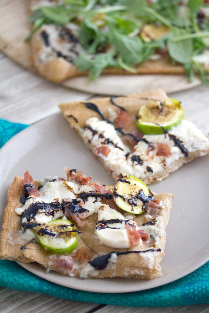 Overhead view of two slices of fig, prosciutto, and ricotta flatbread on a plate with remaining pizza with arugula in background