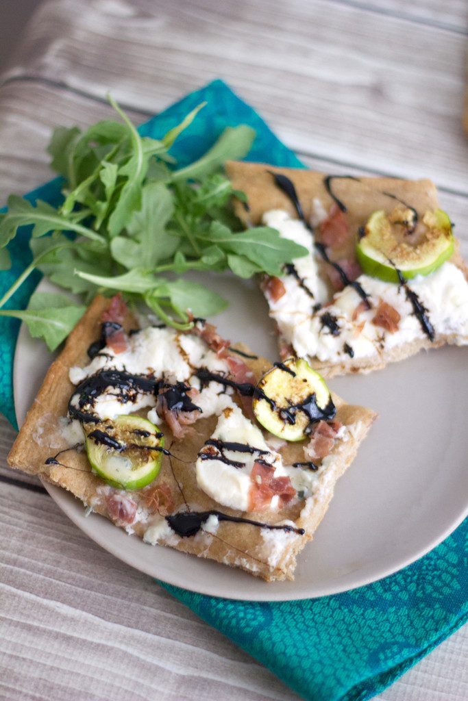 Overhead view of two slices of fig, prosciutto, and ricotta flatbread on a plate with arugula on the side