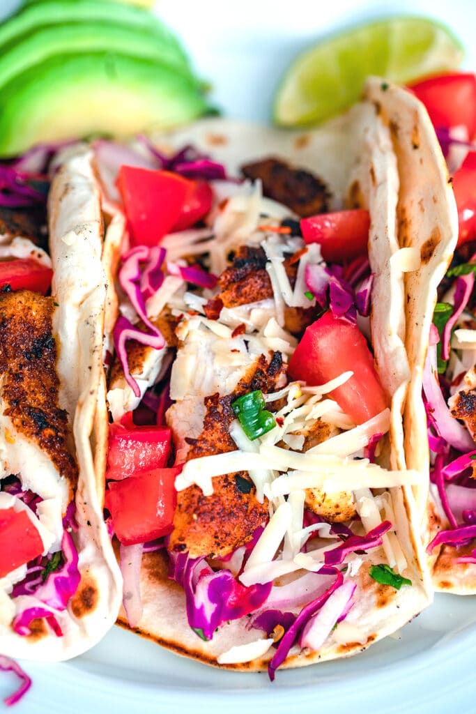 Head-on close up view of a fish taco topped with shredded cheese, tomatoes, and red cabbage with sliced avocado and lime wedge in the background