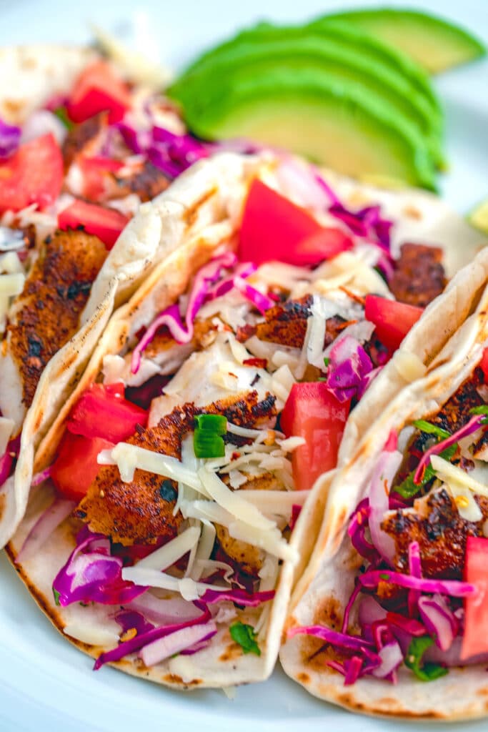 Close-up for fish tacos with bright purple cabbage, red tomatoes, and shredded cheese with sliced avocados in the background