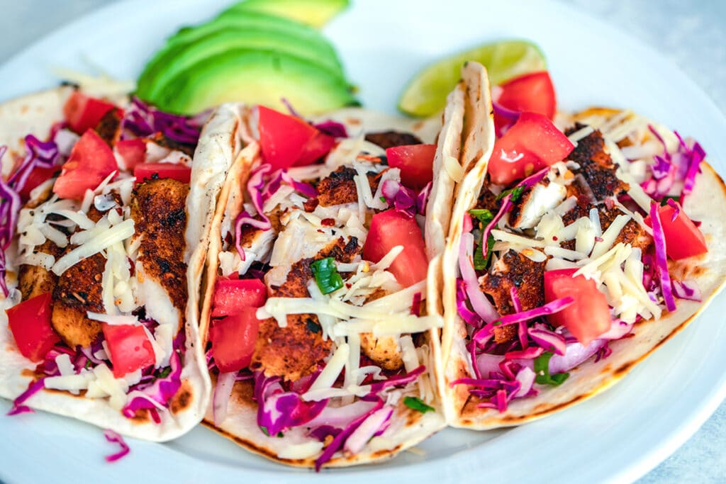 Landscape head-on view of three fish tacos on a white plate topped with red cabbage, tomatoes, and shredded cheese with sliced avocado and a lime wedge in the background