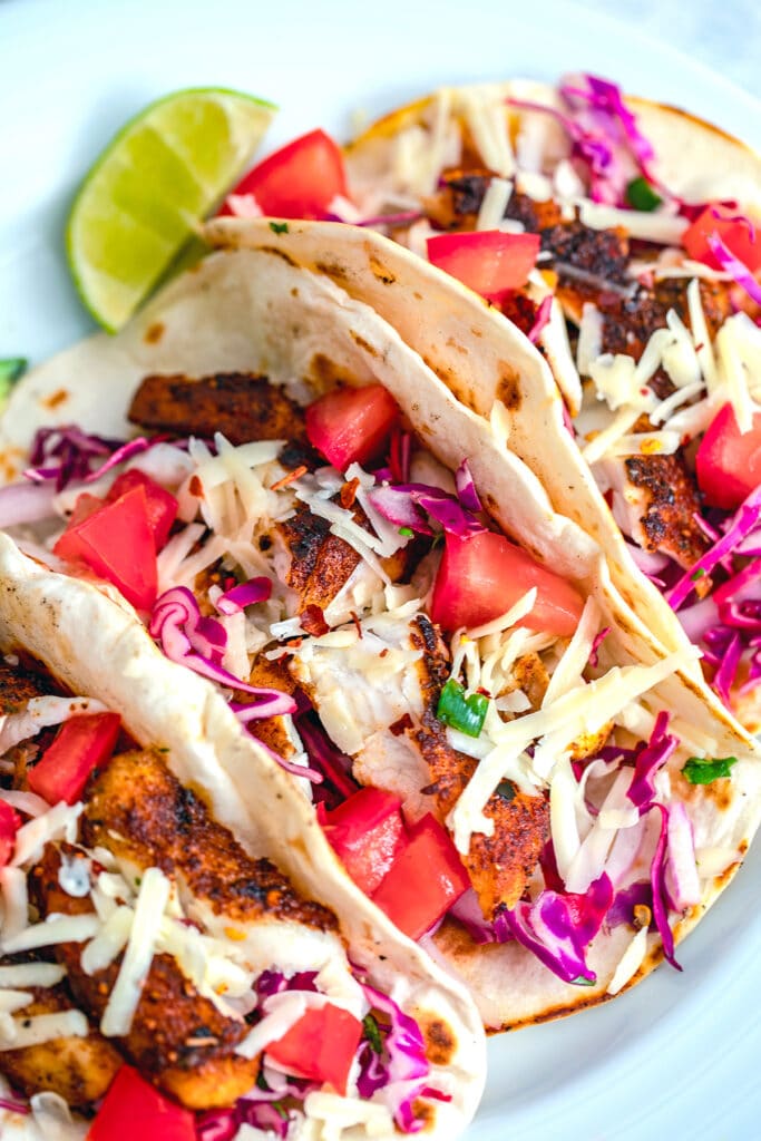 Overhead view of three fish tacos topped with shredded cheese, red cabbage, and tomatoes and a lime wedge on the side