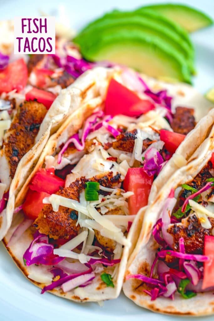 Close-up for fish tacos with bright purple cabbage, red tomatoes, and shredded cheese with sliced avocados in the background and "Fish Tacos" text at the top of the photo