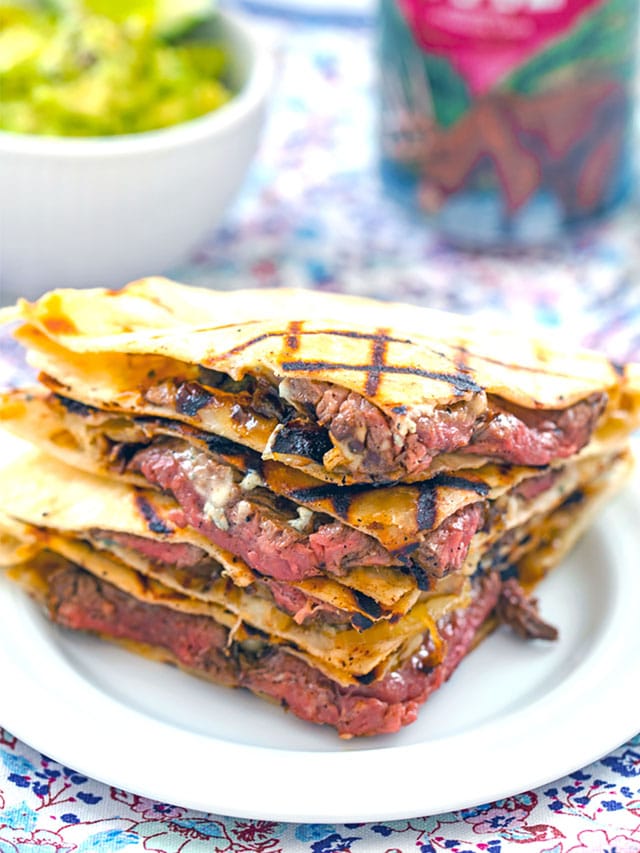 Head-on view of flank steak quesadillas with gorgonzola stacked on each other with bowl of mashed avocado and beer can in the background