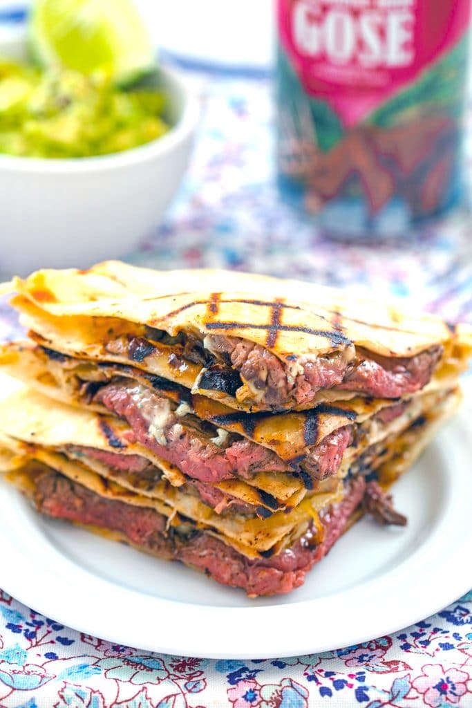 Head-on view of flank steak quesadillas with gorgonzola stacked on each other with bowl of mashed avocado and beer can in the background