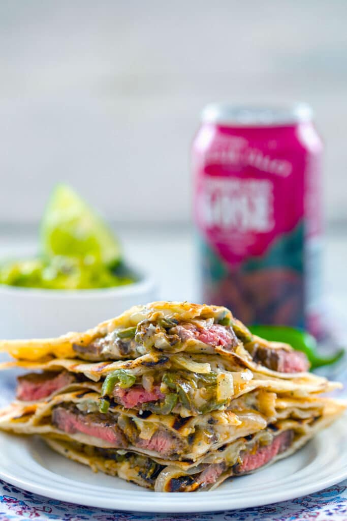 Straight-on view of sliced flank steak quesadillas stacked on top of each other with onions, jalapeño peppers, and gorgonzola cheese with bowl of mashed avocado and beer can in the background