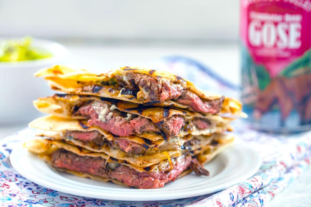 Landscape head-on view of sliced flank steak and gorgonzola quesadillas with onions and jalapeño peppers on a white plate with bowl of mashed avocado and can of beer in background