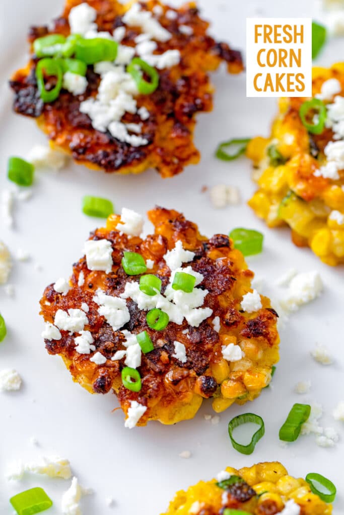 Overhead close-up view of a fresh corn cake on a white platter topped with crumbled feta cheese with scallions and other corn cakes around and recipe title at top