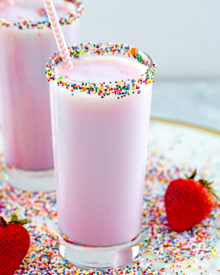 Head-on view of a glass of pink strawberry milk with sprinkle rim with sprinkles and strawberries all around and second glass of milk in background