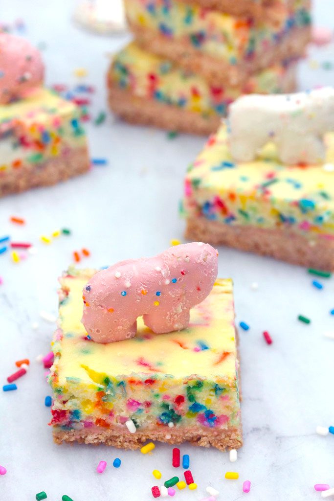 Head-on view of frosted animal cracker cheesecake bar with pink animal cracker on top, lots more cheesecake bars in background, and rainbow sprinkles scattered around