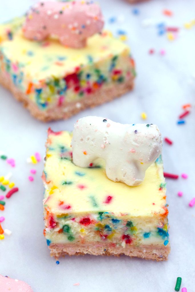 Closeup of a frosted animal cheesecake bar with white animal cracker on top with second cheesecake bar in background and sprinkles scattered around