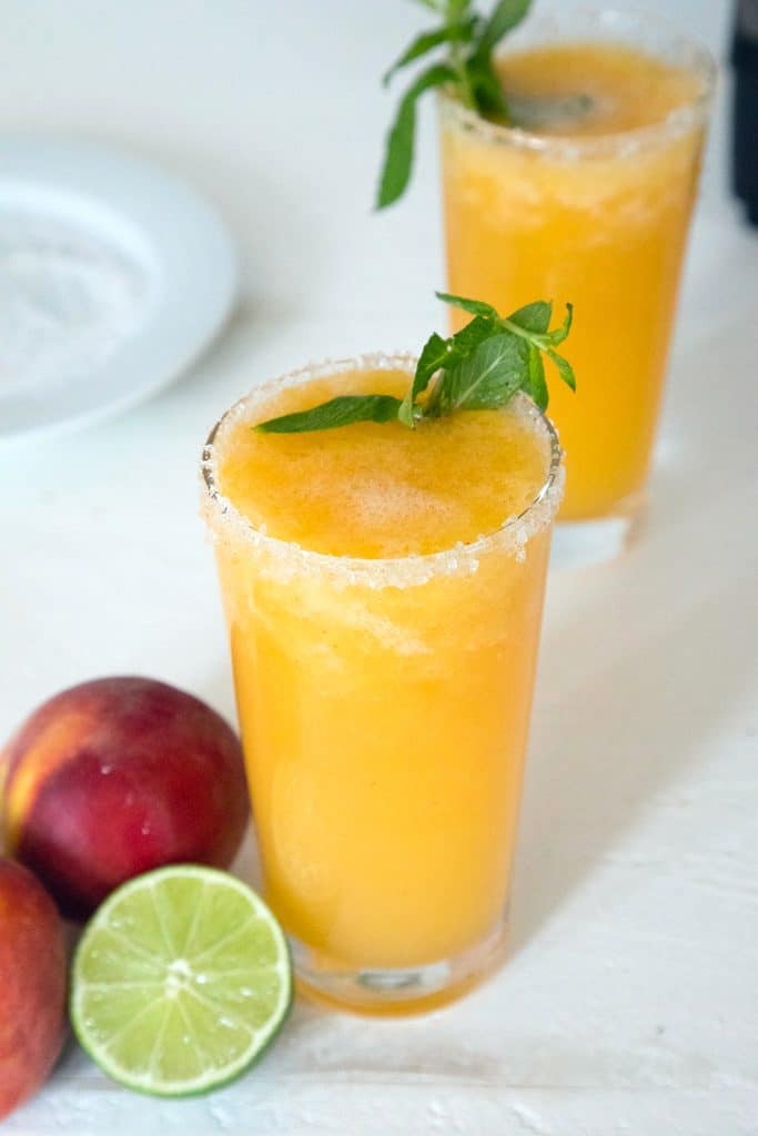 Overhead view of two glasses of bright orange frozen peach margarita topped with mint with a lime half, peaches, and a plate of salt in the background