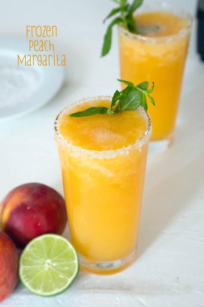 Overhead view of two glasses of bright orange frozen peach margarita topped with mint with a lime half, peaches, and a plate of salt in the background with recipe title at top