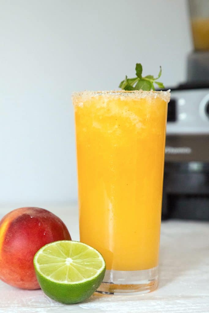 Head-on view of a tall bright orange glass or frozen peach margarita with a lime half and peach in front of it and a Vitamix blender in back