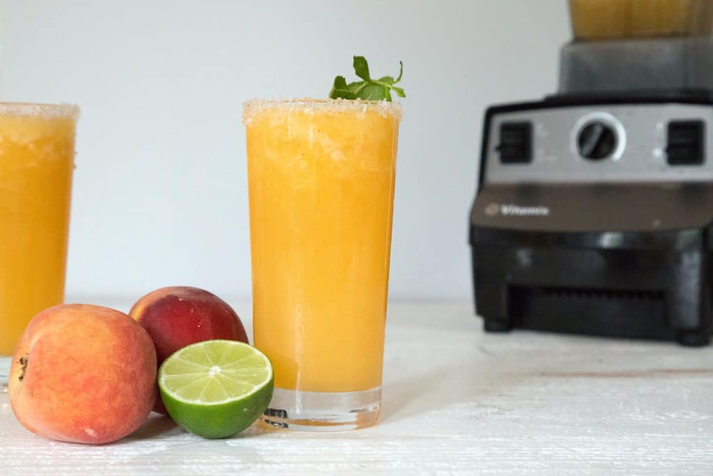 Landscape view photo of a tall glass of bright orange frozen peach margarita with a lime half, peaches, second margarita, and Vitamix blender in background