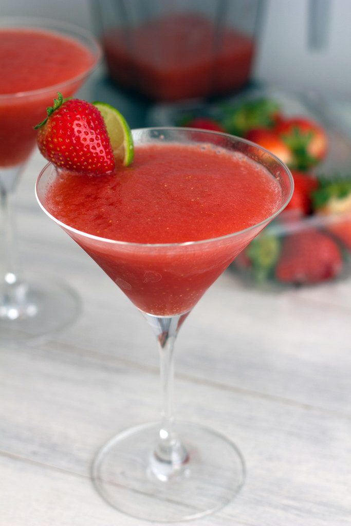 Head-on view of frozen strawberry cosmo with strawberry lime garnish, another cosmo and strawberries in the background