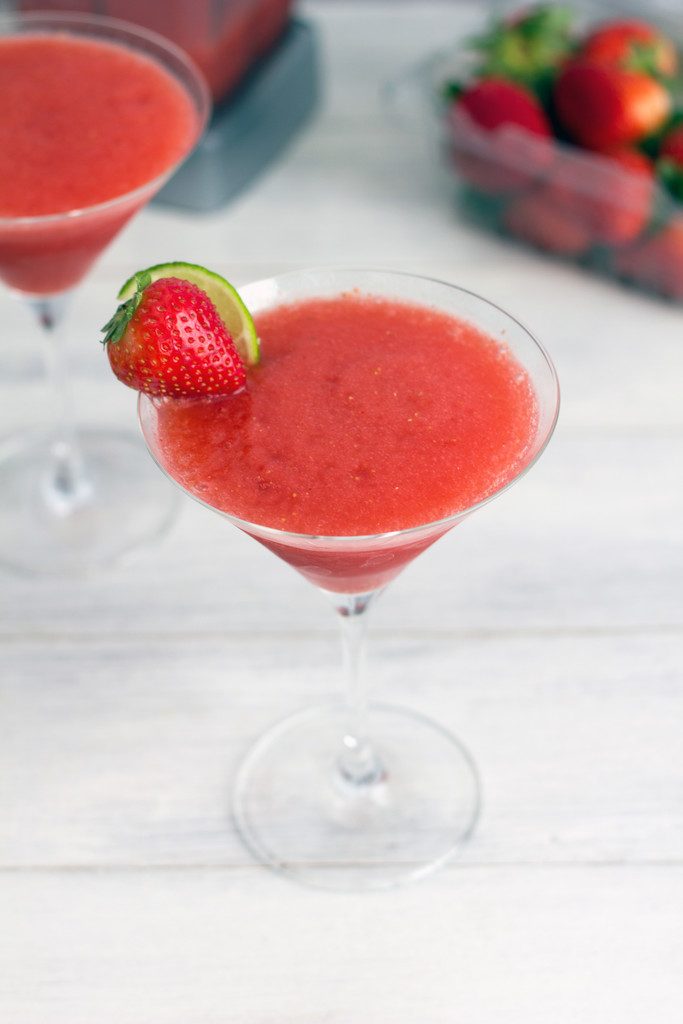 Overhead view of frozen strawberry cosmo in a martini glass with strawberry lime garnish and second glass, strawberries, and blender in the background