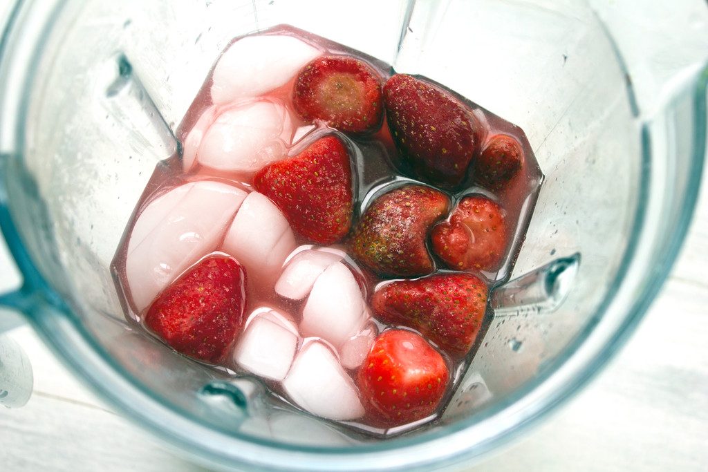 Overhead view of blender filled with ice, cranberry juice, lime juice, vodka, and frozen strawberries for frozen strawberry cosmos