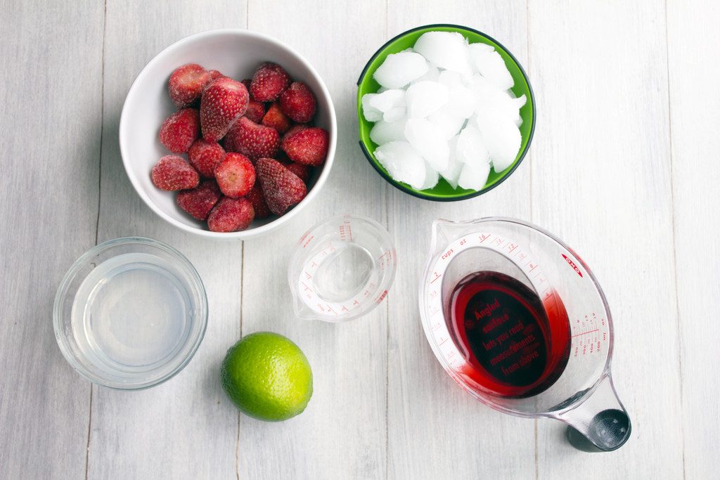 Flat lay of ingredients for frozen strawberry cosmo, including frozen strawberries, cranberry juice, vodka, lime, and ice