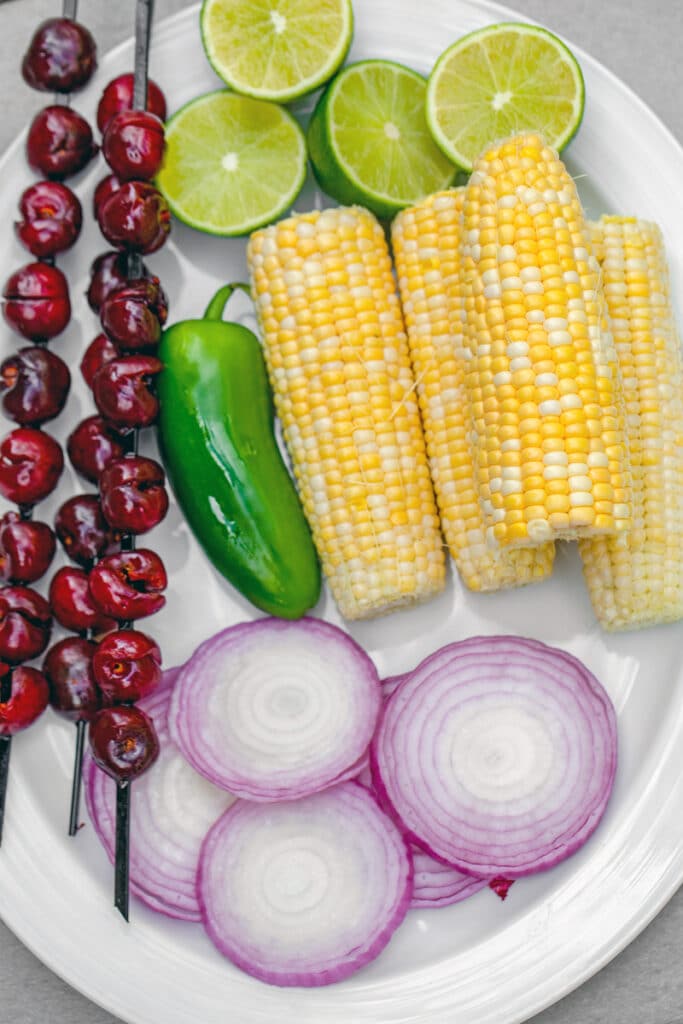 Overhead view of platter with corn on the cob, jalapeño, cherries on skewers, lime halves, and sliced red onions